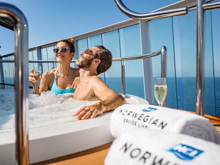Mann und Frau entspannen in einem Whirlpool auf einem Kreuzfahrtschiff mit Meerblick und einem Handtuch mit der Aufschrift 'NORWEGIAN CRUISE LINE'