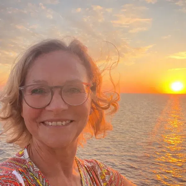 Frau mit kurz geschnittenem Haar trägt ein gemustertes Oberteil und steht vor einem Sonnenuntergang am Meer