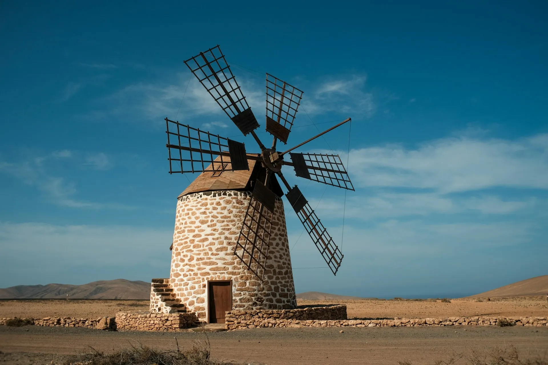 Steinerne Windmühle mit sechs Flügeln in karger Landschaft unter blauem Himmel