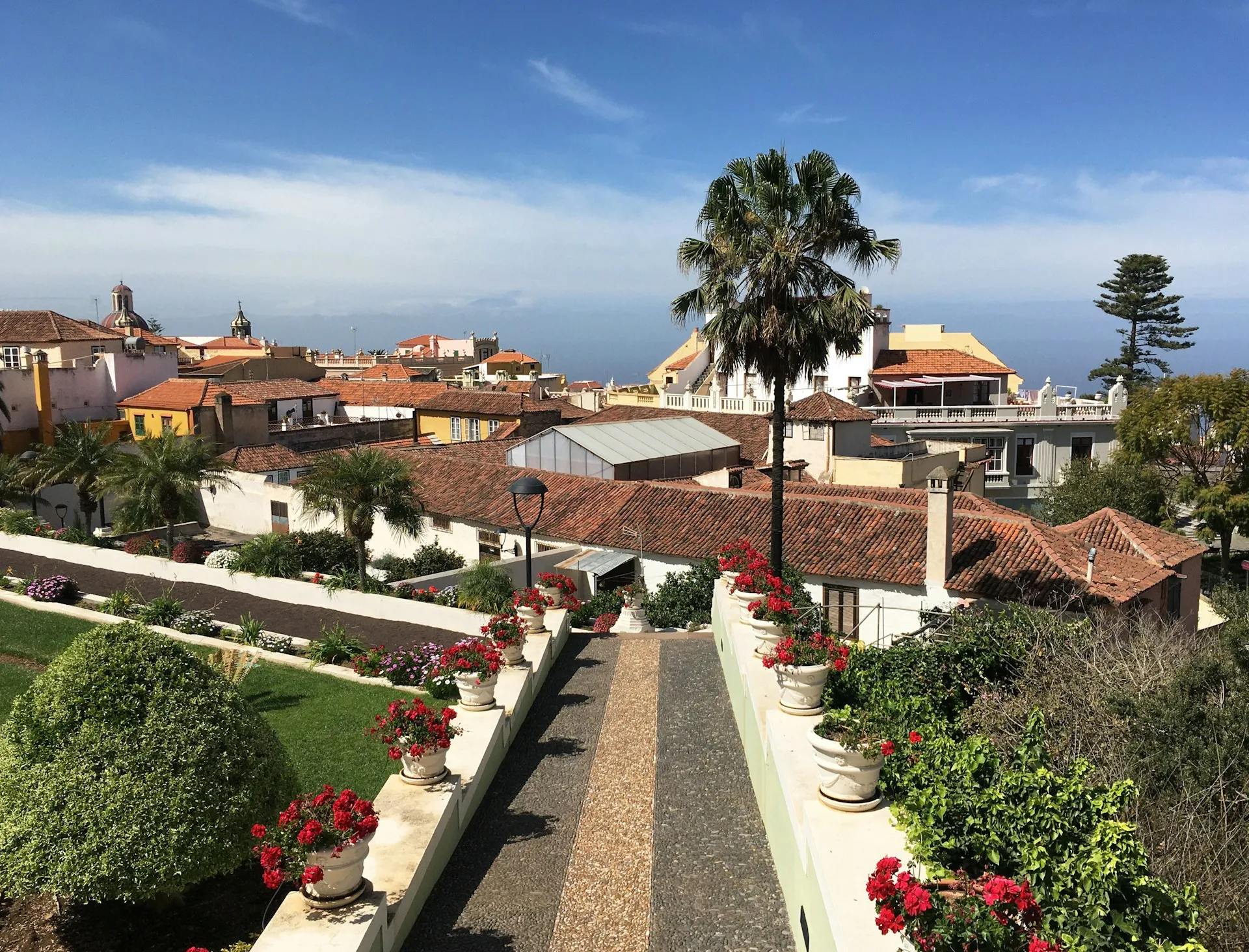 Blick über einen Gartenweg mit Blumentöpfen und roten Blumen zu Häusern mit Ziegeldächern und Palmen im Hintergrund