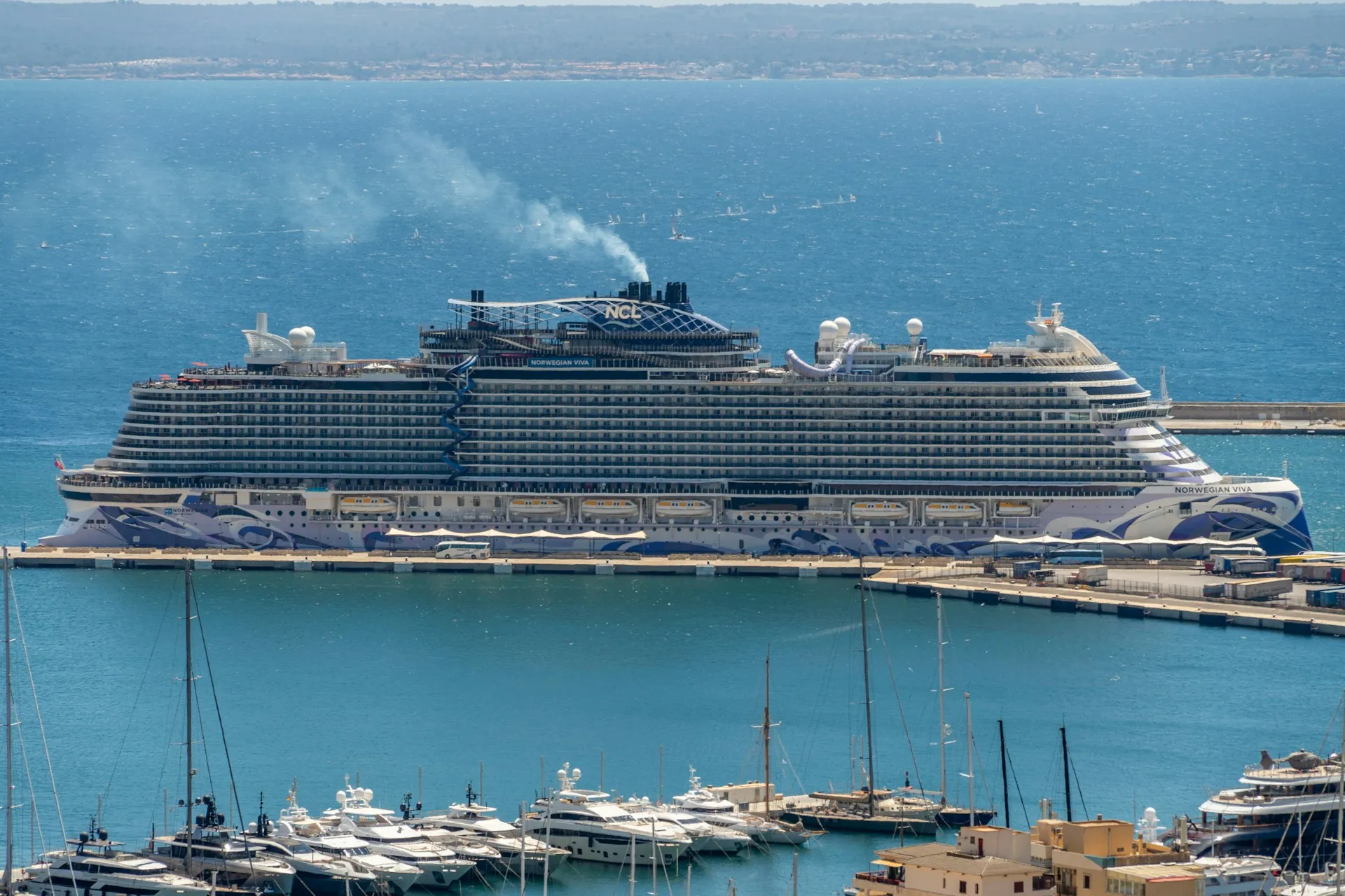 Felsklippe mit Häusern am Wasser und mehreren Booten darunter sowie einem großen Kreuzfahrtschiff auf dem Meer unter blauem Himmel
