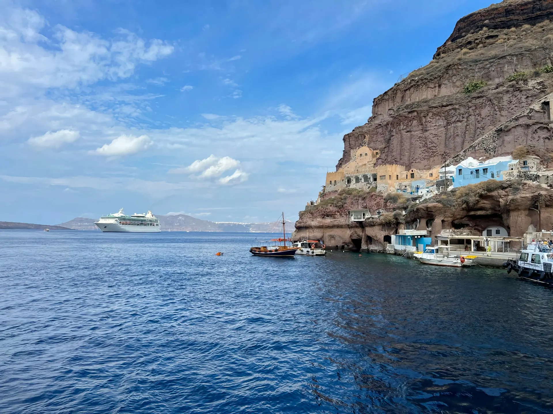 Felsklippe mit Häusern am Wasser und mehreren Booten darunter sowie einem großen Kreuzfahrtschiff auf dem Meer unter blauem Himmel