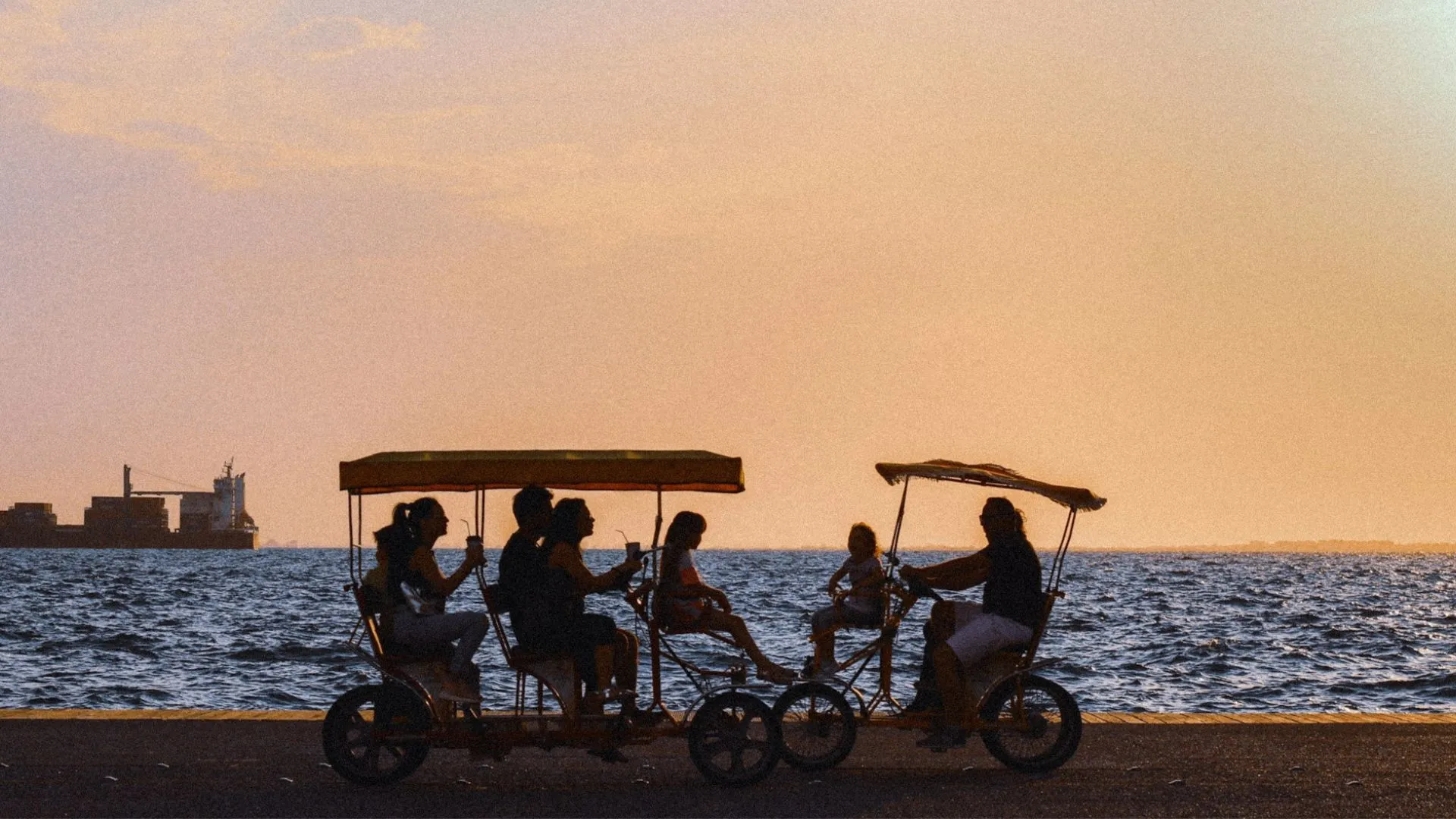 Silhouetten von mehreren Personen auf zwei vierrädrigen Fahrrädern mit Sonnendach am Wasser bei Sonnenuntergang