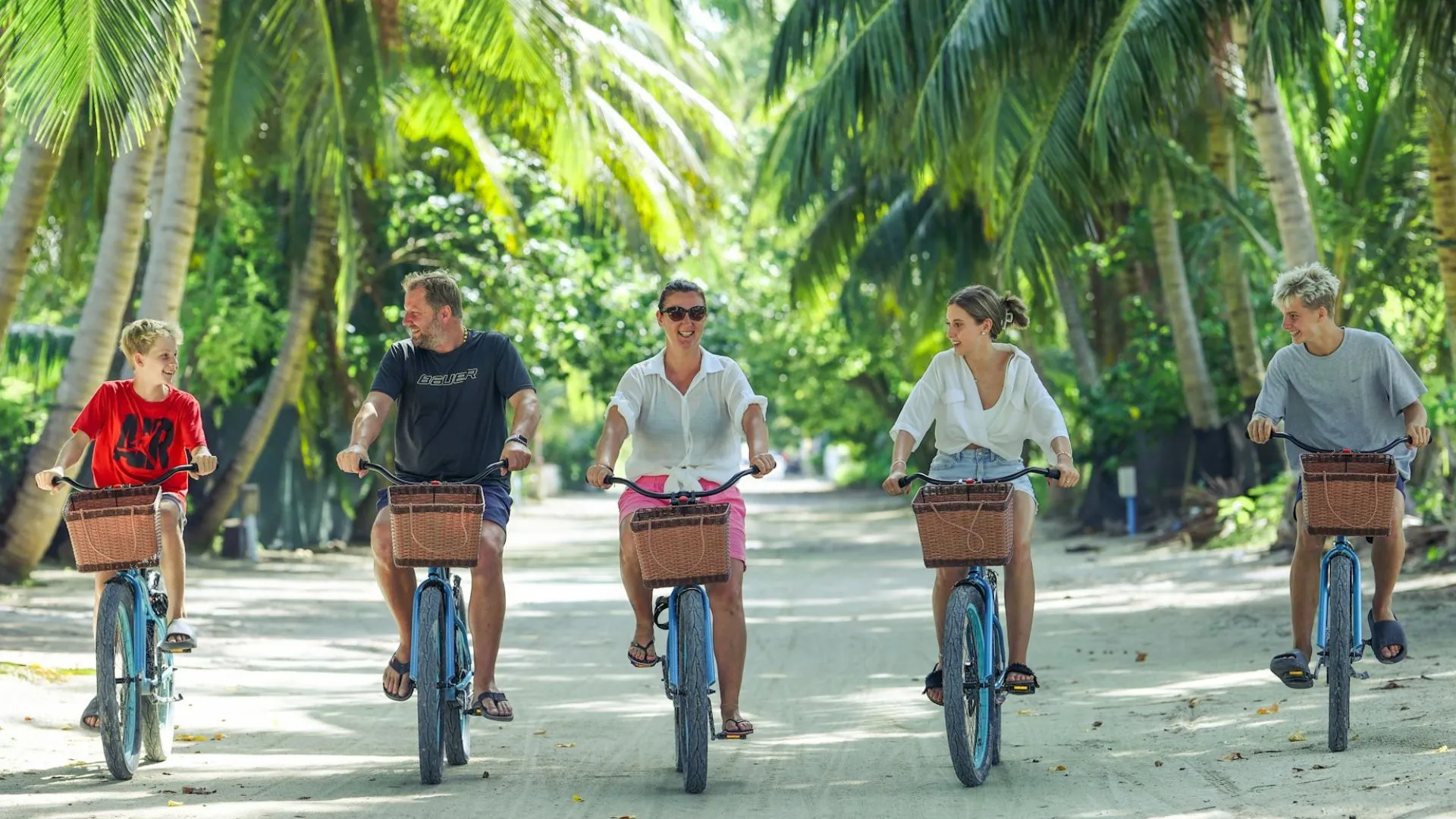 Fünf Personen fahren auf Fahrrädern mit Körben auf einem von Palmen gesäumten Weg in tropischer Umgebung