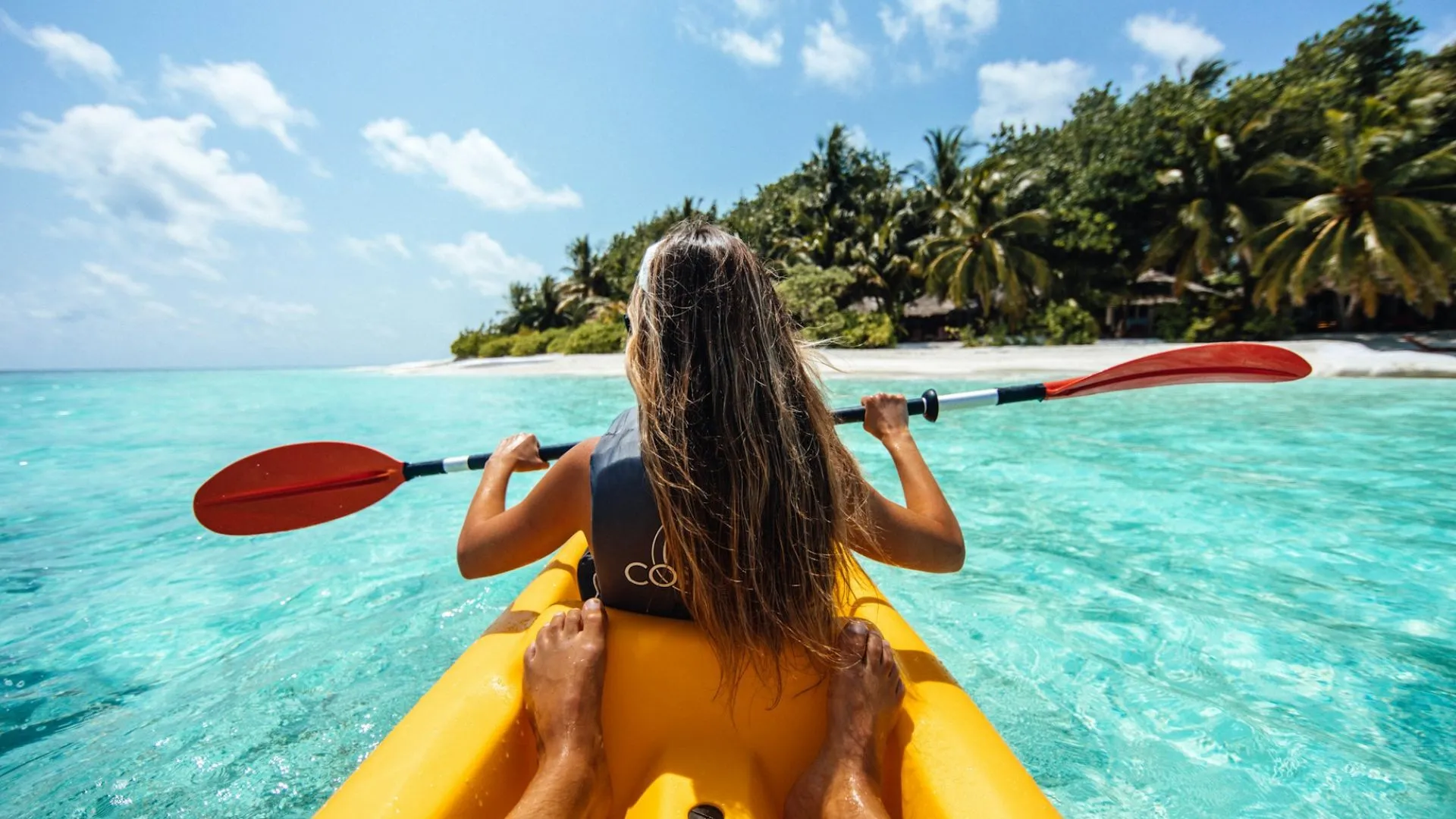Person mit langen Haaren sitzt in einem gelben Kajak auf türkisfarbenem Wasser und hält ein Paddel, Blick auf eine tropische Insel mit Palmen
