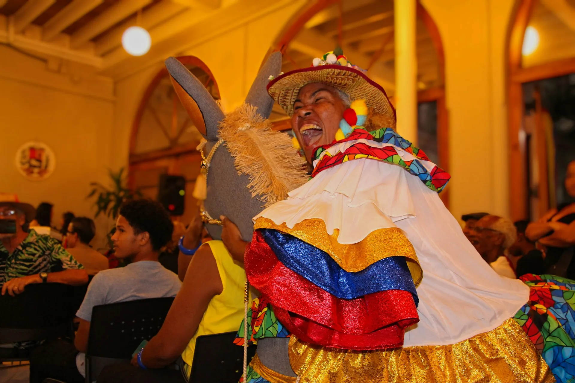 Person in buntem traditionellem Kostüm mit Hut sitzt auf einem Esel in einem Raum mit Holzbalkendecke und mehreren Zuschauern