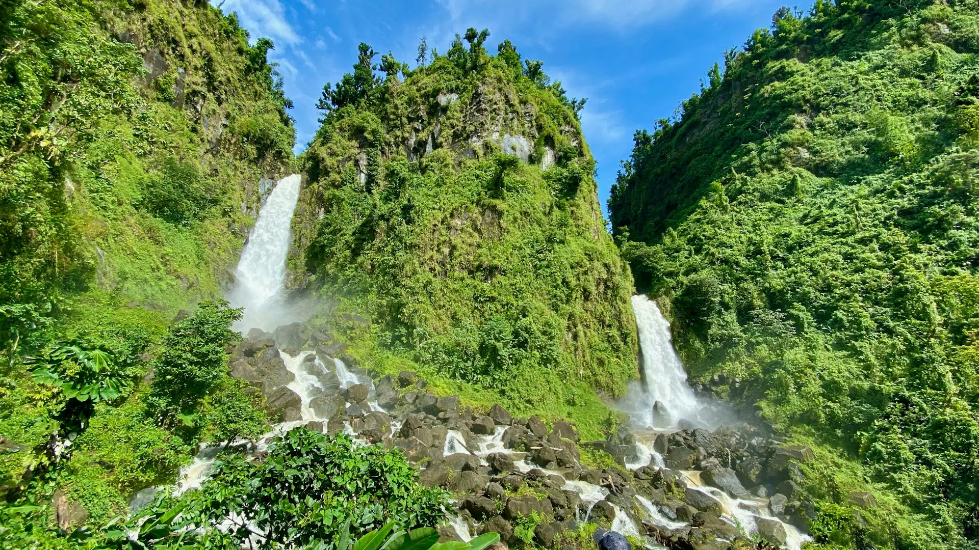 Zwei Wasserfälle stürzen zwischen bewachsenen Felsen in einen felsigen Flusslauf unter blauem Himmel
