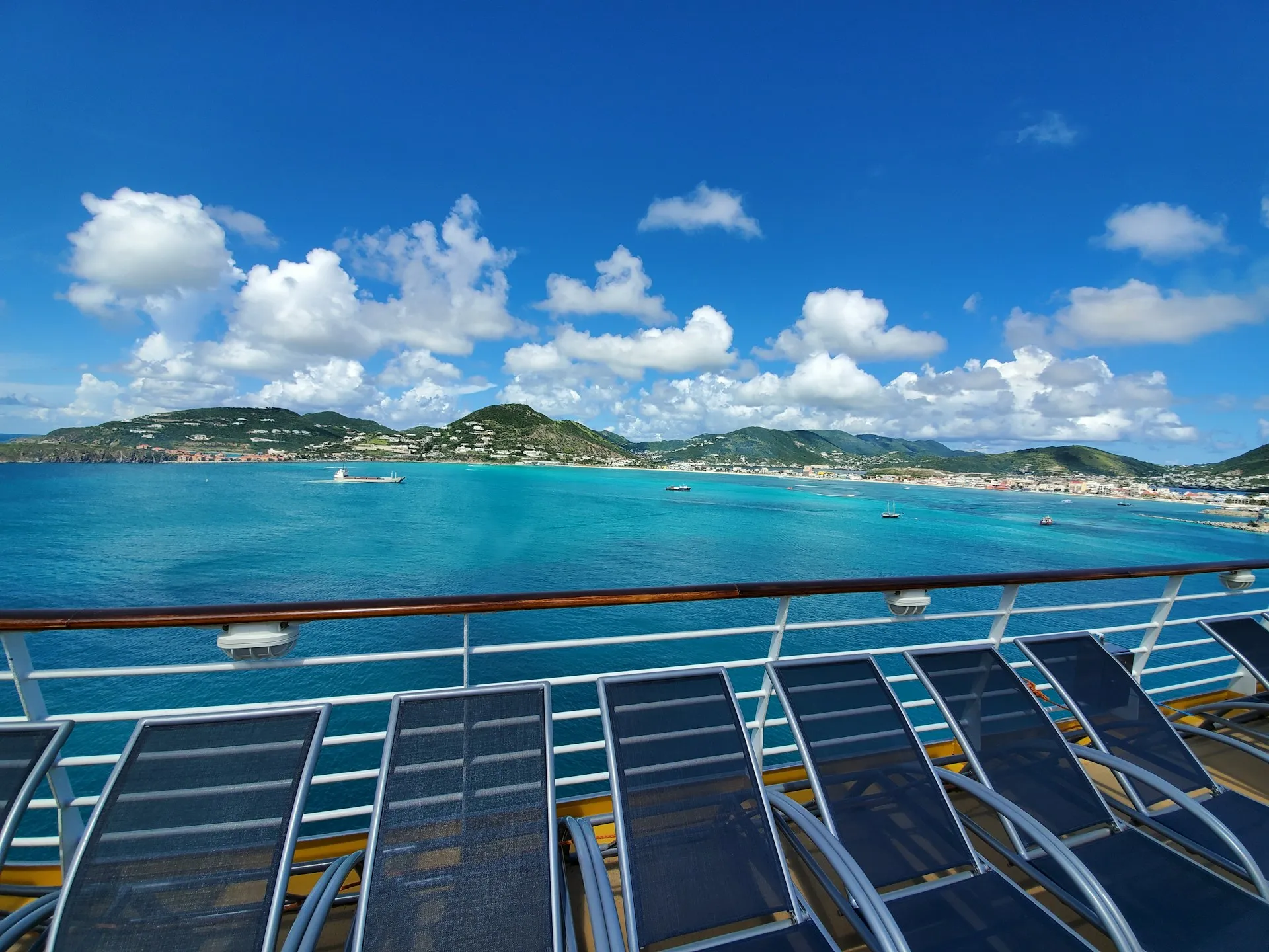 Blick von einem Schiff mit Liegestühlen auf das Meer und eine hügelige Küstenlandschaft unter blauem Himmel mit Wolken