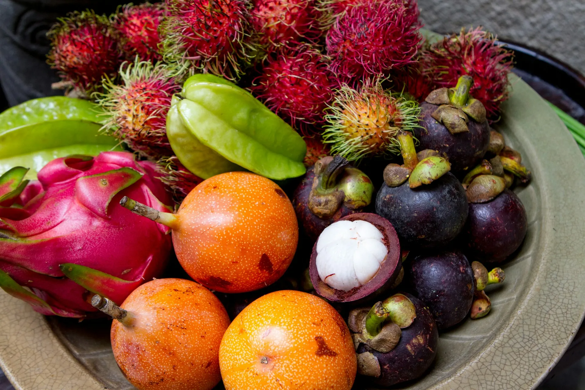 Teller mit verschiedenen exotischen Früchten wie Drachenfrucht, Sternfrucht, Rambutan, Granadilla und Mangostan, wobei eine Mangostan-Frucht aufgeschnitten ist und das weiße Fruchtfleisch zeigt