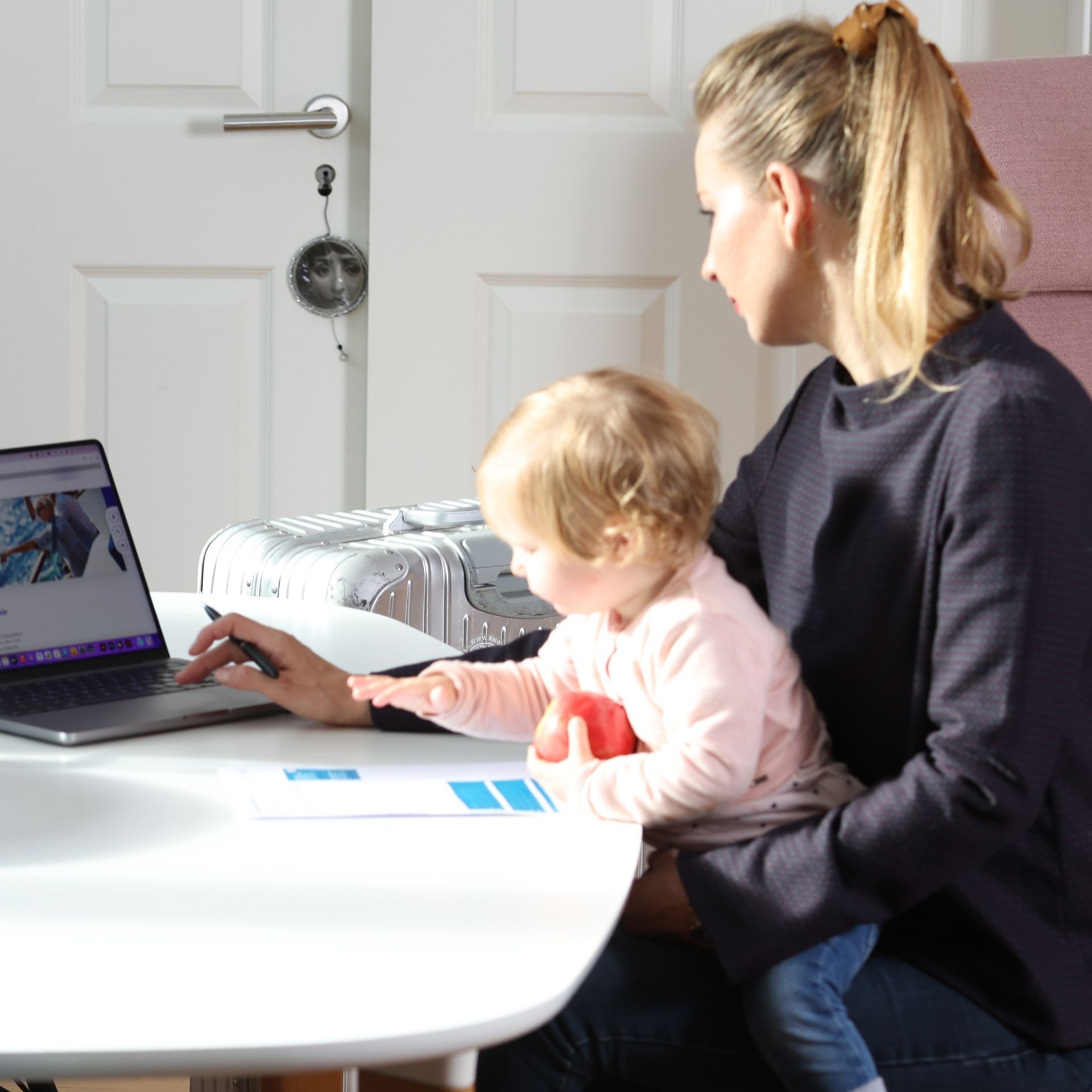 Frau sitzt mit Kleinkind auf dem Schoß an einem weißen Tisch und bedient einen Laptop