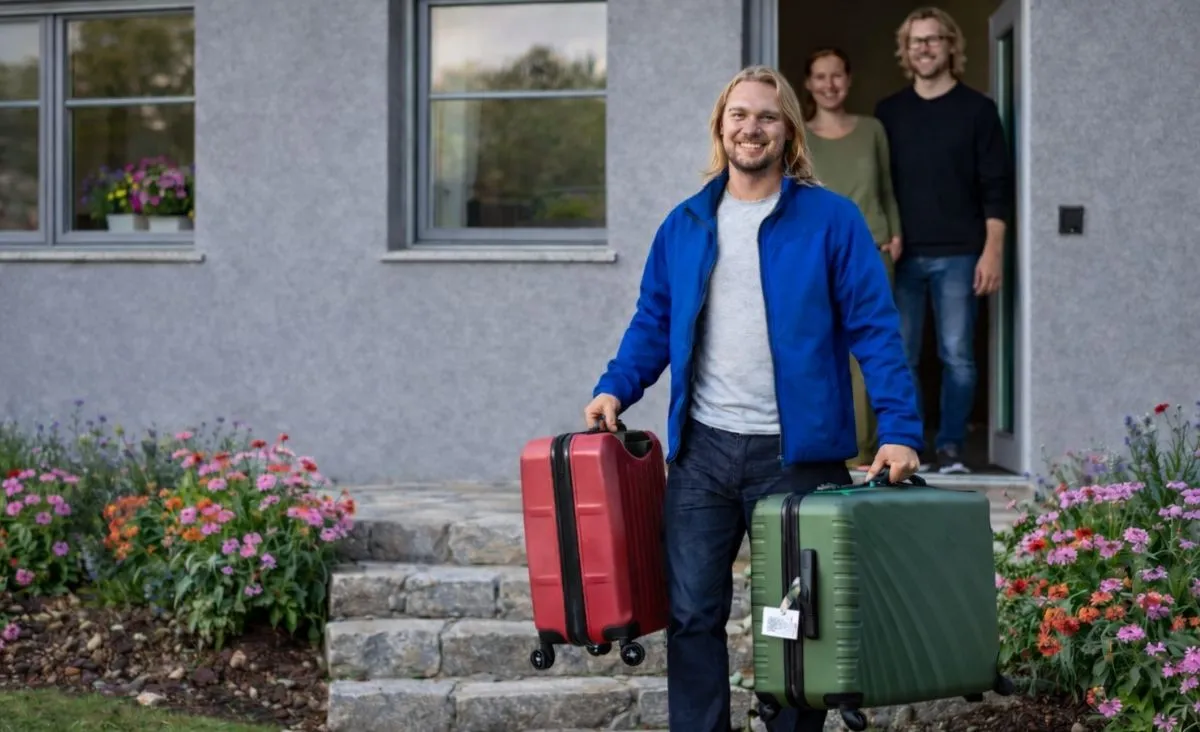 Mann in blauer Jacke trägt zwei Koffer vor einem Haus mit zwei Erwachsenen im Hintergrund