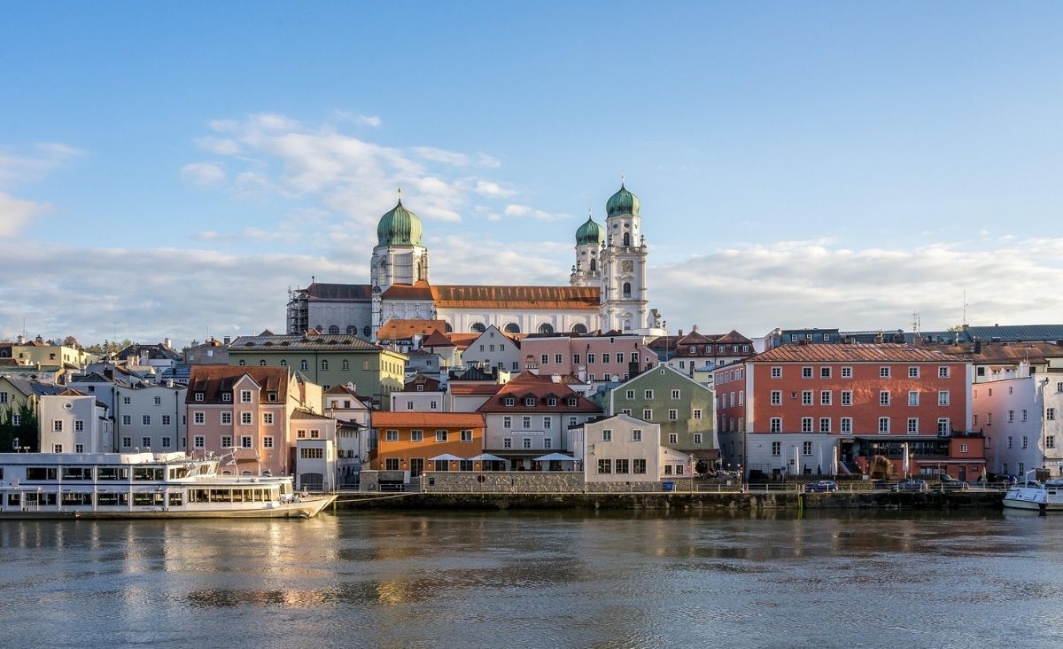 Stadtansicht von Passau mit Fluss im Vordergrund und barocken Gebäuden sowie zwei markanten Kirchtürmen im Hintergrund